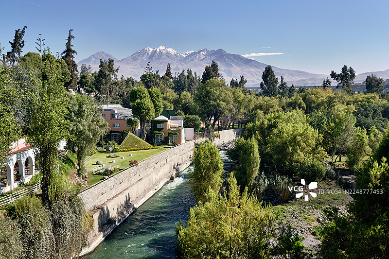 宁静的河流穿过生机勃勃的绿色景观，周围有茂密的树木和石头墙，背景是雄伟的雪山，天空湛蓝，秘鲁阿雷基帕图片素材