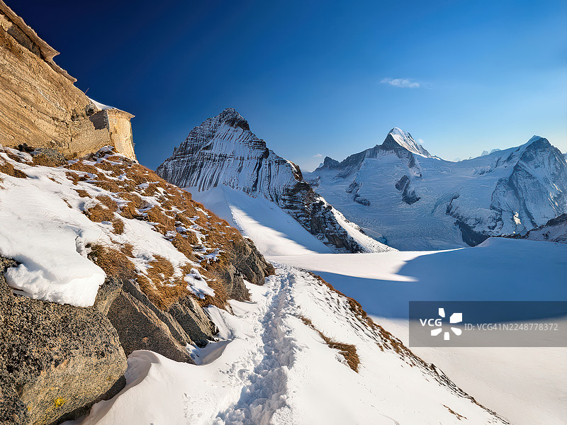 瑞士，小屋：晴朗蓝天下雄伟的雪山，蜿蜒的小路穿过雪景图片素材