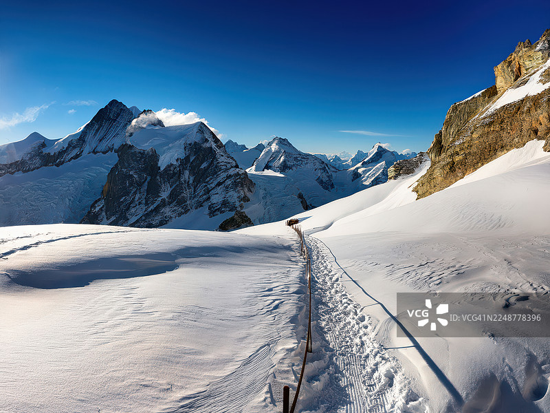 在晴朗的蓝天下，雄伟的雪山，一条蜿蜒的雪路通向远方，瑞士，小屋图片素材