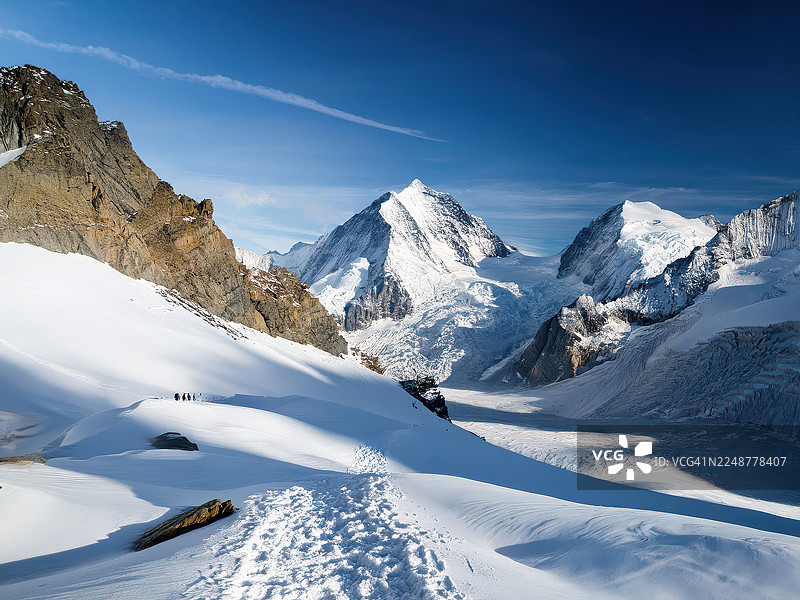 晴朗的蓝天下，雄伟的雪山和冰川，远处雪道上有徒步旅行者，瑞士，小屋图片素材