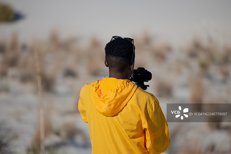 一名男子背影，身穿亮黄色夹克，头戴太阳镜，手持相机，置身于户外，地点为美国阿拉莫戈多。图片素材