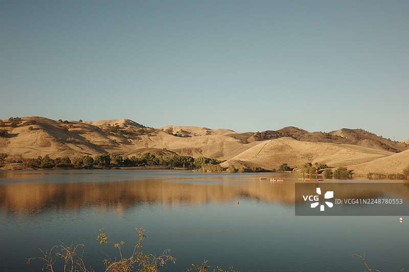 风景优美的宁静湖泊倒映着金色的山丘，在晴朗的蓝天下，营造出宁静和自然之美的感觉，美国，安提阿，康特拉洛马水库图片素材