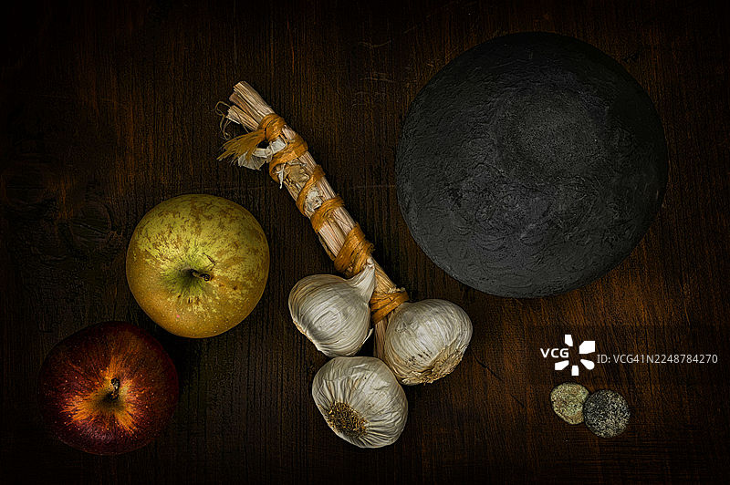 俯视角度拍摄的苹果、大蒜和一个深色纹理球体放在质朴的木桌上，营造出一种黑暗、忧郁的氛围图片素材