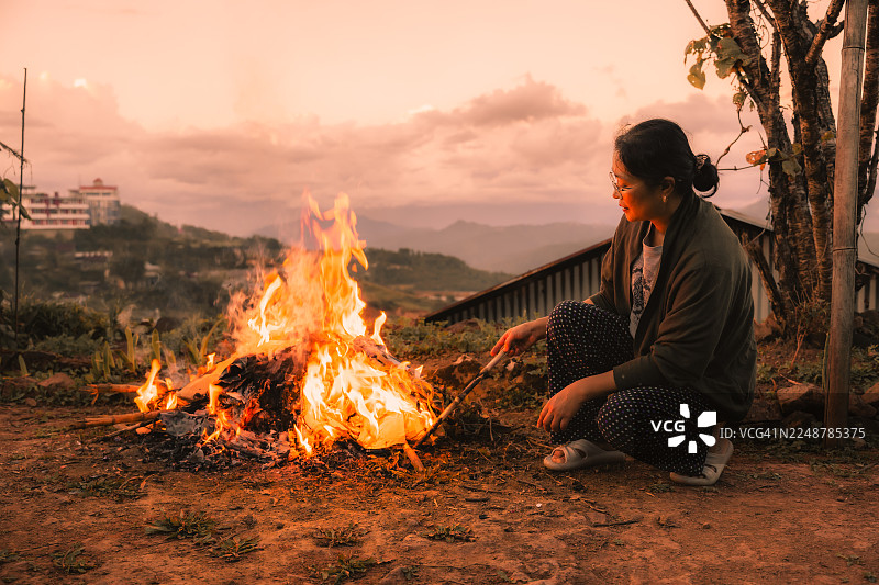 日落时分，一位女性蹲坐在温暖的篝火旁，在山间享受宁静的户外氛围。图片素材