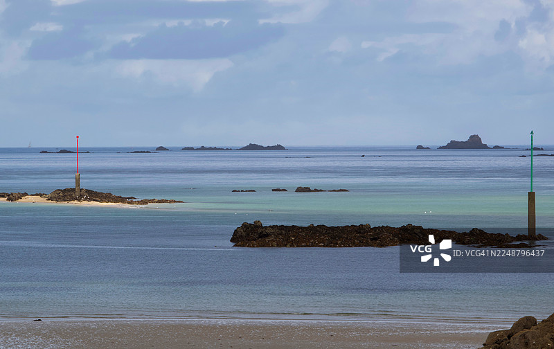 法国迪纳尔，宁静的海滨风光，沙滩延伸至平静湛蓝的海水，零星散布着几座小岩岛，天空布满云彩。图片素材