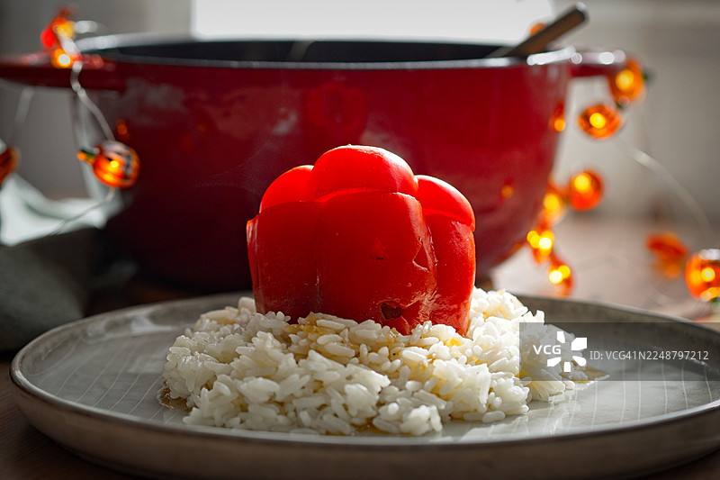 特写：盘子里放着一盘鲜红的酿甜椒，底下铺着白米饭，背景是红色的锅和装饰灯。图片素材