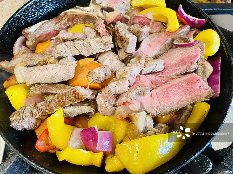 俯视视角，铸铁锅中滋滋作响的牛排、彩椒和洋葱正在烹饪，展示出一顿色彩鲜艳、美味诱人的餐食准备过程图片素材