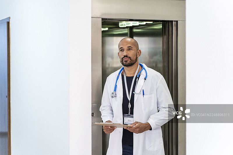 一名男医生，身着白大褂，佩戴听诊器，手持病历夹，正走出医院的电梯图片素材