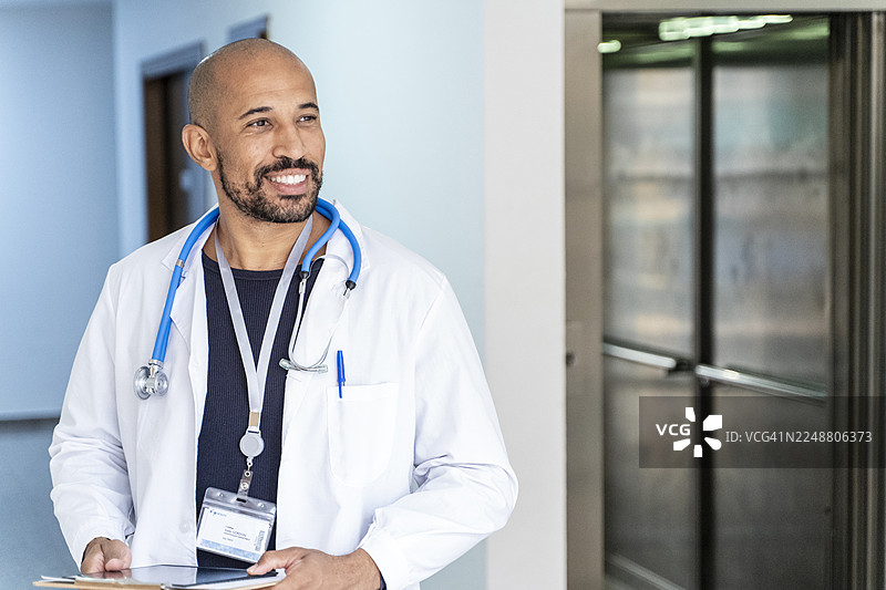 一名身穿白大褂、佩戴听诊器的男医生，手持平板电脑，在医院走廊里微笑着。图片素材