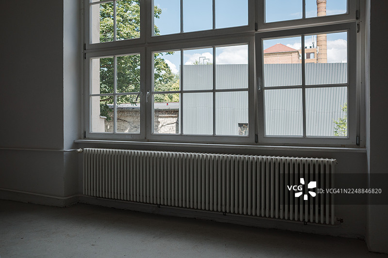 空房间的内部，上方是一扇带有多扇窗格的大窗户，下方是长条形的白色散热器，窗外是绿树成荫的景色。图片素材