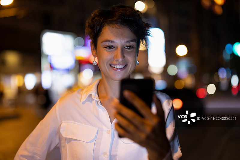 夜晚，一位年轻女子在充满活力的城市街道上，面带微笑地使用智能手机。图片素材