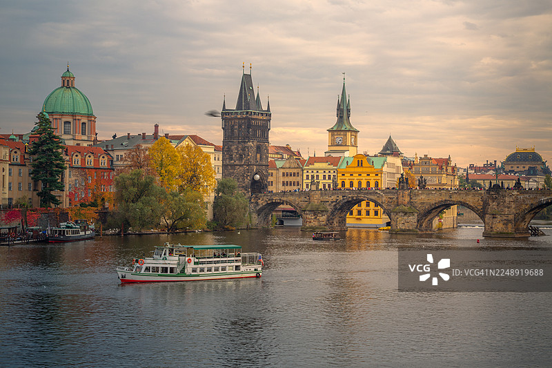 布拉格城市景观，历史悠久的查理大桥横跨伏尔塔瓦河，河上有老城桥塔和色彩缤纷的建筑，在温暖的秋日天空中，一艘旅游船正在河上航行。图片素材