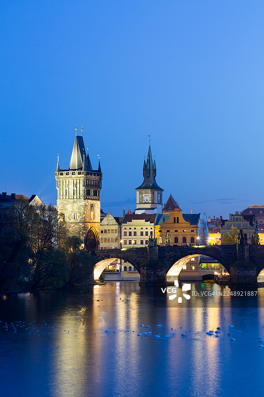 查理大桥在蓝色时刻，历史建筑倒映在伏尔塔瓦河上，白天鹅在灯火辉煌的塔楼附近游弋，这是布拉格日落后著名的地标。图片素材