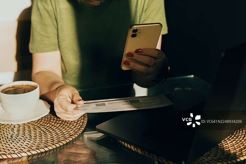 在咖啡馆的藤编桌上，一个人一边喝着咖啡，一边使用智能手机，旁边放着收据和笔记本电脑图片素材