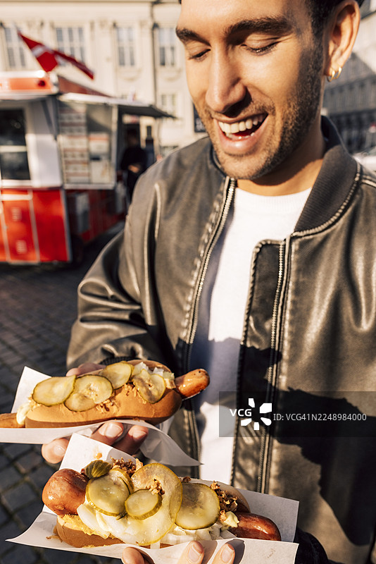年轻男子站在街头，手持新鲜热狗，面带微笑图片素材