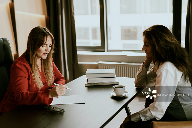 在明亮的办公室里，两位女士在会议期间于办公桌旁讨论文件图片素材