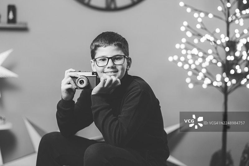 一个快乐的男孩带着相机，在有圣诞树灯光的节日家庭环境中图片素材