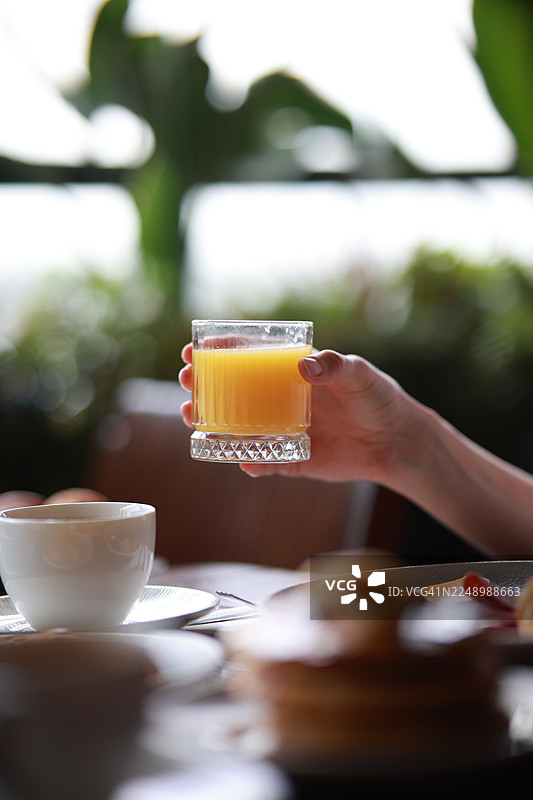 特写镜头，一只手在传统的土耳其早餐中拿着一杯新鲜的橙汁，柔和的自然光照耀，背景是模糊的绿色植物，体现了早晨的热情好客和健康的生活方式，适用于食品和旅行设计。图片素材