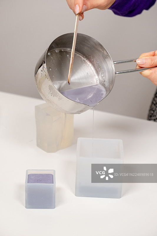 从不锈钢锅中将融化的紫色肥皂混合物倒入硅胶模具的特写镜头，在室内手工工作坊中拍摄，俯视构图，突出纹理、着色剂使用以及手工设计DIY护肤品的概念图片素材
