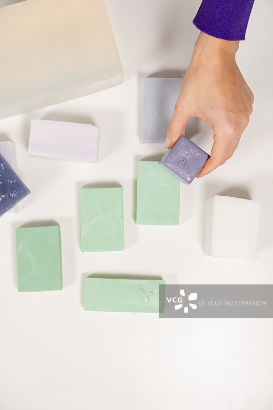 干净工作台面上，多块手工制作的长方形肥皂成品中，一人正在挑选彩色手工皂的俯视图，DIY手工艺环境，突出美容和健康设计的艺术护肤品生产图片素材