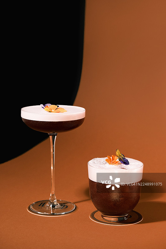 两杯深紫色鸡尾酒的正面视图，顶部有浓密的泡沫，并饰有可食用的花朵，背景为温暖的棕色工作室。优雅的混合饮品展示，搭配对比鲜明的玻璃器皿，现代调酒理念，适用于酒吧菜单和品牌视觉形象。图片素材