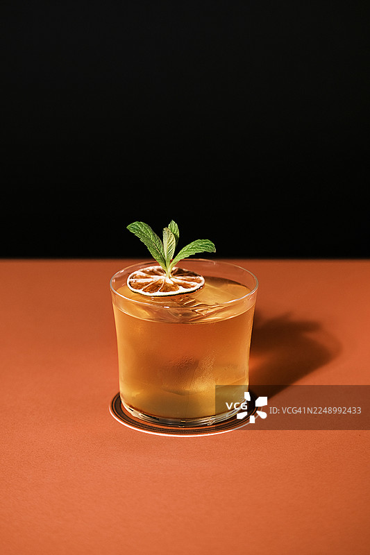 低球杯中琥珀色鸡尾酒的特写，配有薄荷和干柑橘装饰，温暖色调的台面上极简的影棚布置，现代饮品呈现概念，用于菜单设计和品牌推广。图片素材