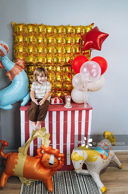 快乐的二岁生日！可爱的幼儿男孩坐在梳妆台上，背景是装饰精美的气球拍照区。图片素材