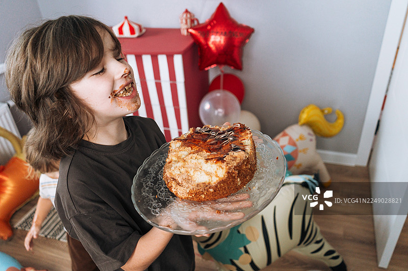 一个七岁的男孩在生日派对上，用牙齿咬着生日蛋糕，房间里装饰着节日气氛，充满童趣。图片素材