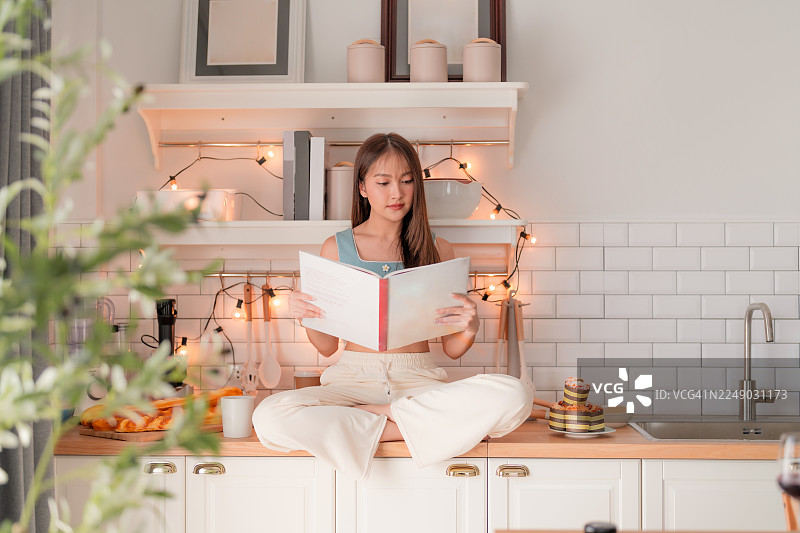 年轻女子在家中享受悠闲放松的时刻，舒适地坐在厨房柜台上，全神贯注地阅读一本好书图片素材
