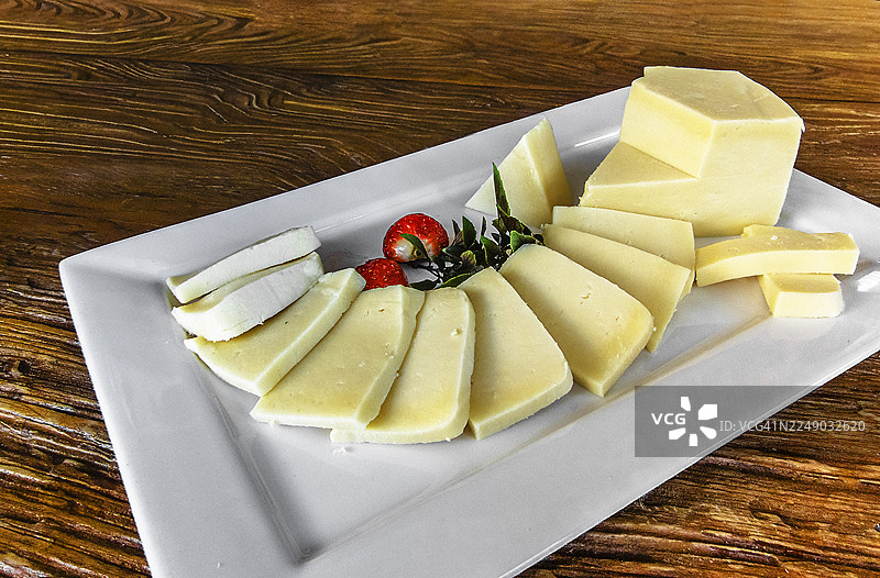 白盘上摆放着帕玛森干酪、布里干酪和陈年奶酪等多种奶酪，并用草莓和绿叶装饰。背景为木质表面。图片素材