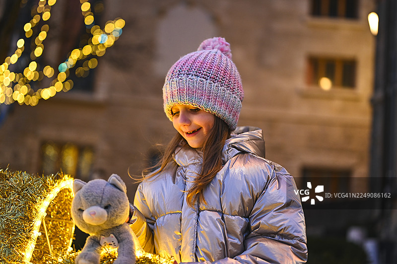 女孩在冬夜里抱着玩具猫，背景是闪烁的圣诞灯光图片素材