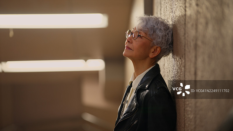 一位年长的女性斜靠在墙壁上，抬头望向城市图片素材