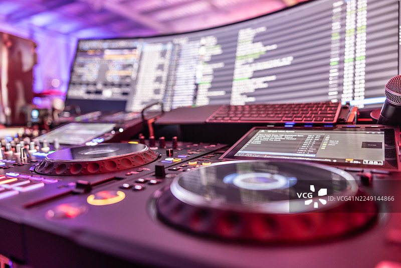 一个先进的DJ设备，景深吸引着人们的注意力聚焦在前景的金属搓盘细节上。背景是一个巨大的弧形屏幕，深色背景上显示着白色文字，使整个场景充满了高科技音乐氛围。图片素材