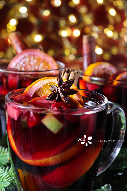特写镜头，三杯装有香料和水果的热葡萄酒，配有肉桂棒、八角、切块的苹果、橙子片，以及圣诞装饰品、人造云杉针小枝，背景有闪烁的彩灯，焦点在前景。图片素材