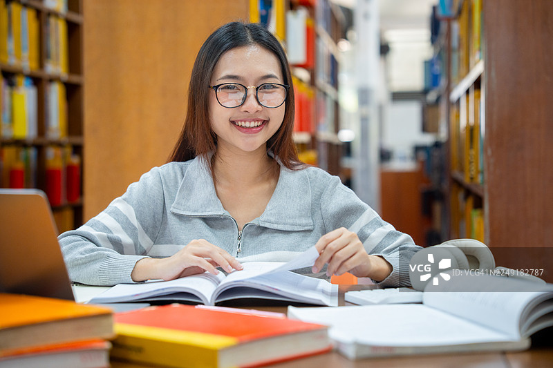 教育与学校概念。年轻女子在图书馆读书。美丽的亚洲女大学生微笑着，手里拿着书，坐在图书馆里。学习与教育图片素材