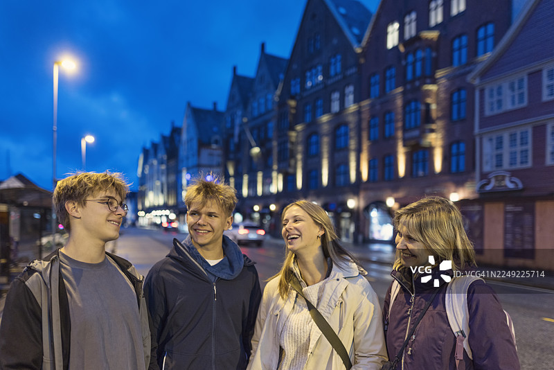 夜幕下，母亲和青少年们站在卑尔根的布吕根海滨，身后是历史悠久的神圣罗马帝国汉萨同盟建筑和街道灯光。图片素材
