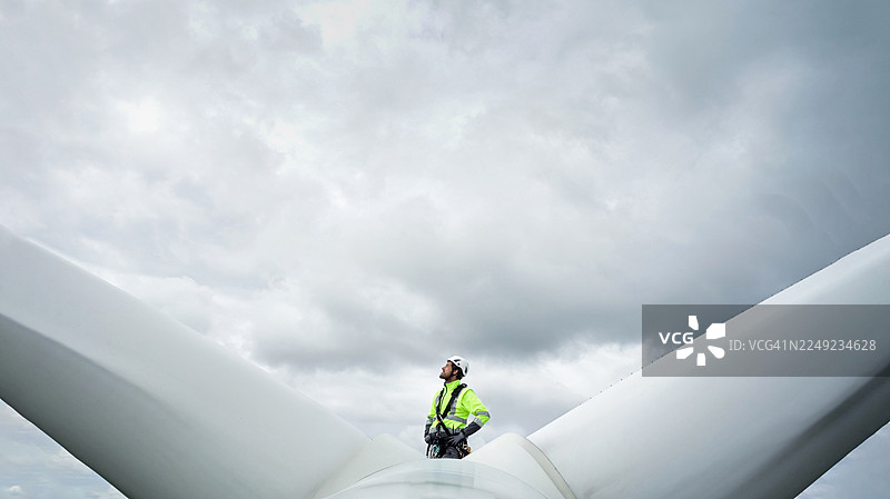 专业的绳索技术员，检查员站在风力涡轮机的轮毂（顶部）上，位于叶片之间并向上看。背景是戏剧性的天空。双手叉腰。图片素材