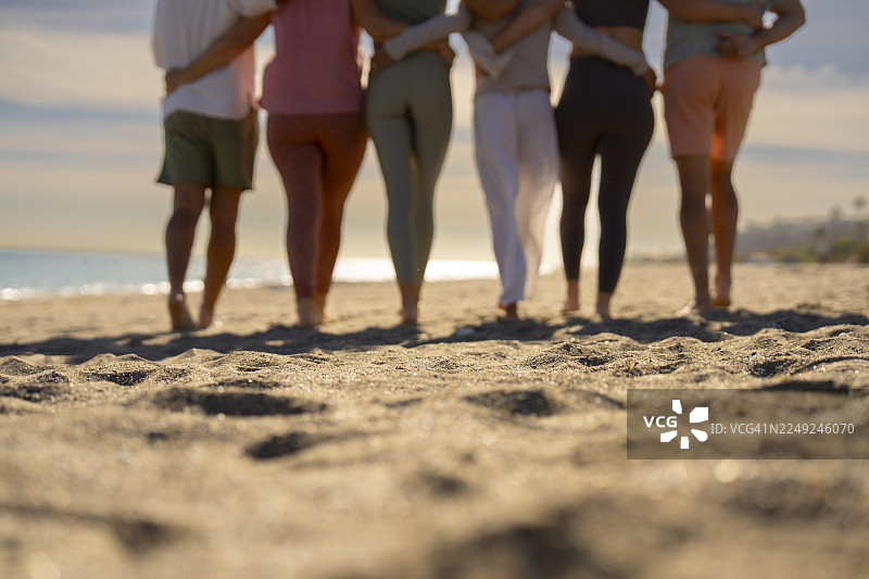 一群朋友在日落时分于海滩上并肩行走并拥抱，从后面拍摄图片素材
