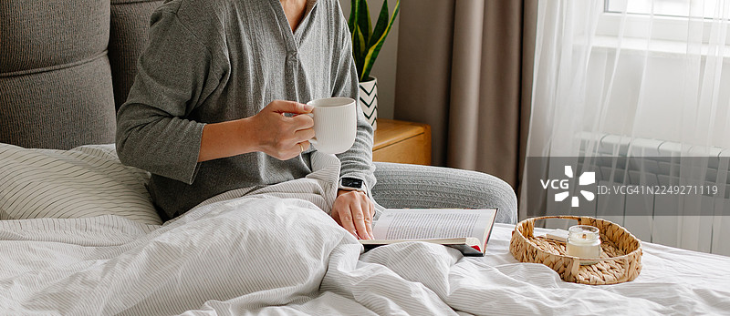 舒适的床上早晨：一个人拿着咖啡杯，旁边点着蜡烛看书图片素材
