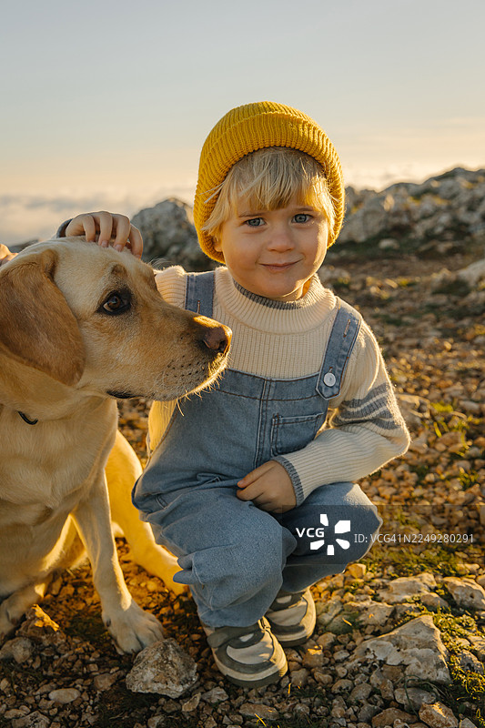 穿着黄色绒帽的小男孩在落日时分与他的狗狗在岩石户外散步时依偎在一起图片素材
