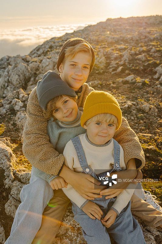 日出时分，三个兄弟姐妹在岩石山上拥抱，金色时光里的温暖与家庭探险图片素材