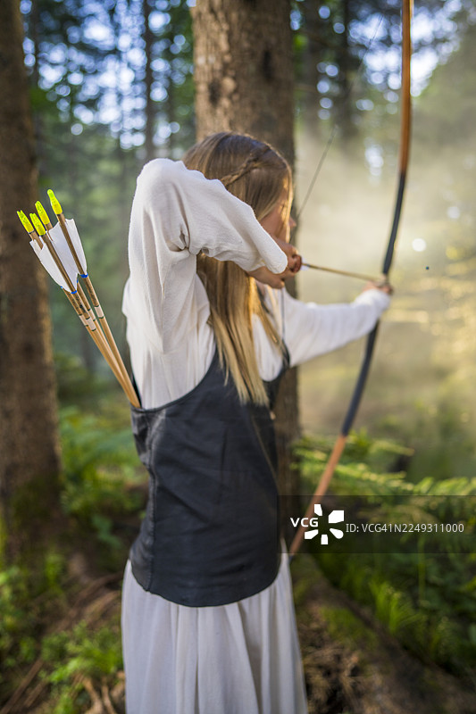 阳光明媚的森林中，年轻的高加索女弓箭手正在娴熟地拉弓。图片素材