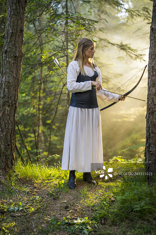 身着中世纪服装的年轻白人女性弓箭手在阳光明媚的森林中练习射箭图片素材