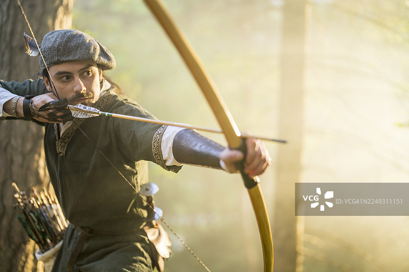 阳光明媚的森林里，一位年轻的高加索男性弓箭手正持弓瞄准图片素材