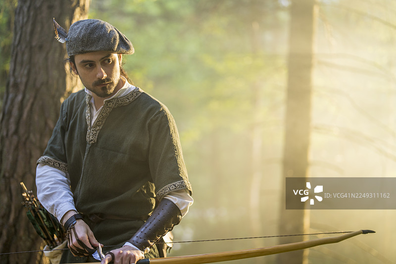 身着中世纪服装的弓箭手在阳光明媚的森林中，弓已拉满，随时准备射击。图片素材