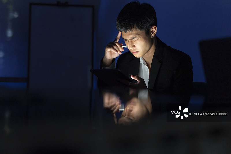 年轻男子在昏暗的办公室加班，同时使用智能手机和数码平板电脑图片素材