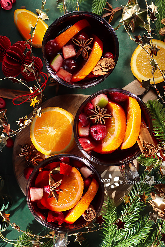 木板上放着三杯热气腾腾的香料水果热红酒，配有肉桂棒、八角、切块的苹果、橙子片、圣诞装饰、人造云杉针枝、金色星星挂饰，背景为绿色，俯视角度。图片素材