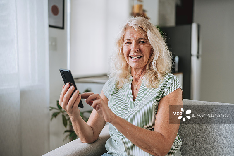 一位快乐的老年女性在家中微笑着使用智能手机，看向镜头。图片素材