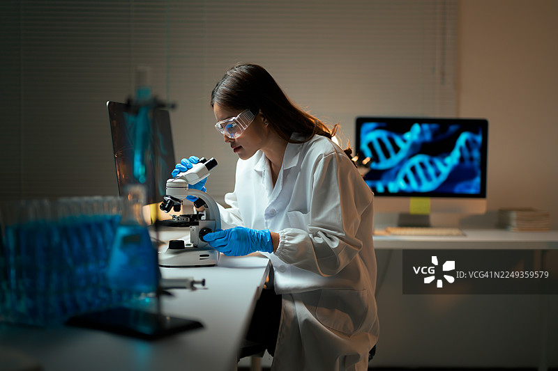 一位女研究员在昏暗的高科技实验室里，使用显微镜检查样本，进行 DNA 和科学分析工作。图片素材