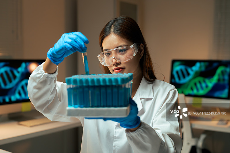 身穿实验服、戴着安全眼镜的女科学家正在研究基因数据，手中拿着装有蓝色液体的试管图片素材